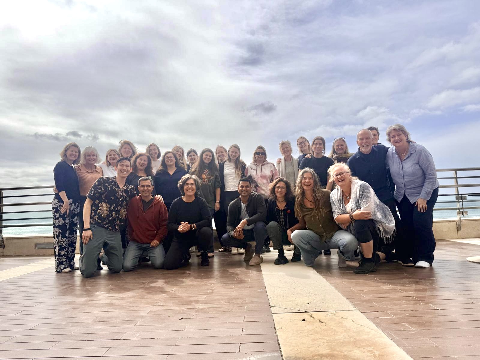 A group posing together on a wooden terrace beside the water under a cloudy sky.