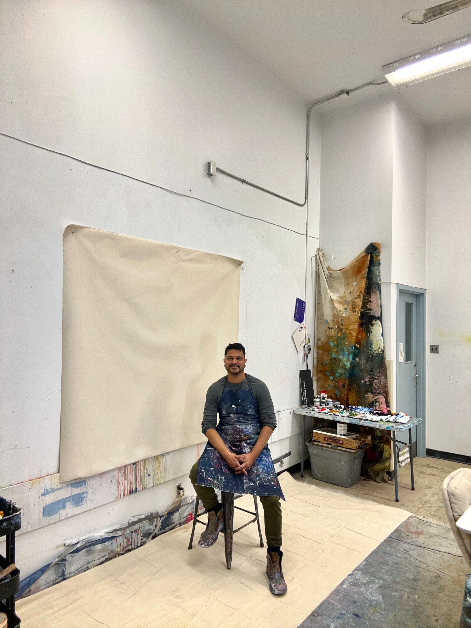 Anand seated in his art studio wearing a paint-covered apron, beside a canvas and painting materials.